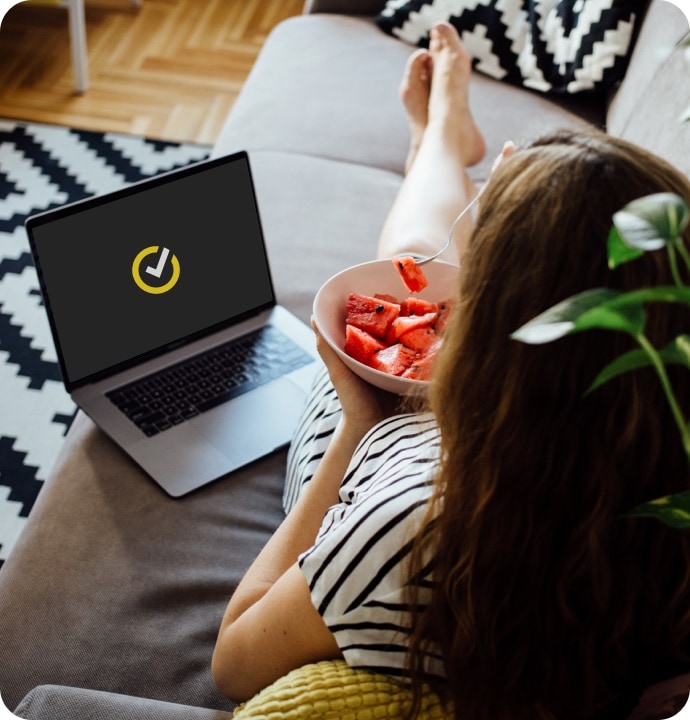 Kvinna som ligger i en soffa och äter frukt från en skål med en bärbar dator bredvid sig med Norton-logotyp