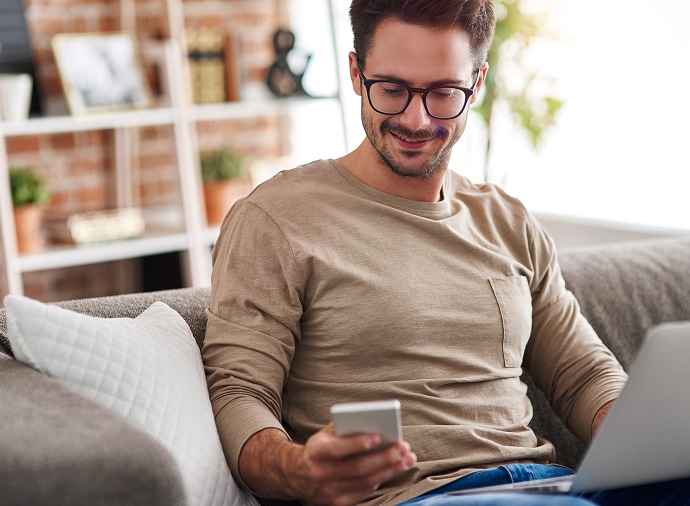 Man som sitter i en soffa och tittar på sin mobil med en laptop i knäet